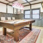 A game room with a pool table on a patterned rug, vintage-style popcorn machine, chairs against the wall, decorative wall paneling, pennants, and a stained-glass light fixture—perfect for luxury athlete accommodations in Lake Arrowhead during the Ho'Omau Altitude Haus training camp.