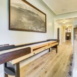 A bright living room inspired by luxury athlete accommodations in Lake Arrowhead during the Ho'Omau Altitude Haus training camp features a shuffleboard table along a light wood floor, a large framed landscape photo, and a comfy white sofa beside built-in shelves with books and decor.