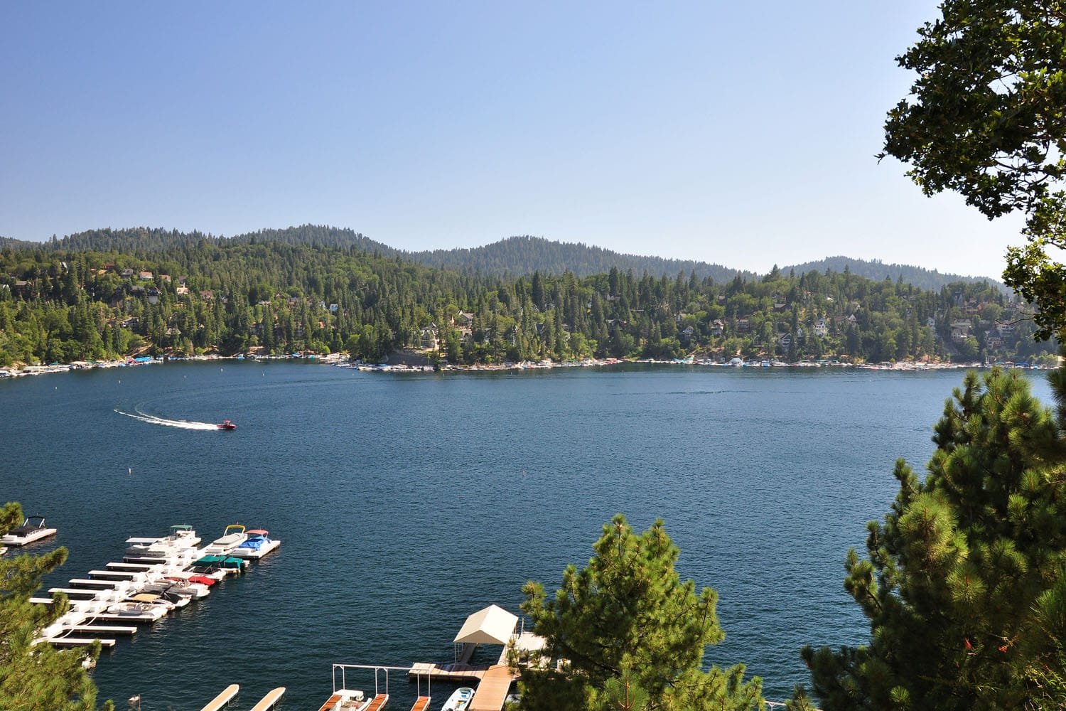 Lake Arrowhead surrounded by forested hills, with boats docked at a marina in the foreground and a small boat moving across the water under a clear blue sky. The perfect setup for the Ho'Omau Altitude Haus multisport training camp.