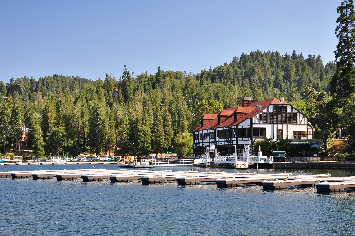 A large Tudor-style building with a red roof sits by the edge of Lake Arrowhead, California, surrounded by forested hills. Several empty docks extend into the calm water under a clear blue sky. An ideal experience for the Ho'Omau Altitude Haus multisport training camp.
