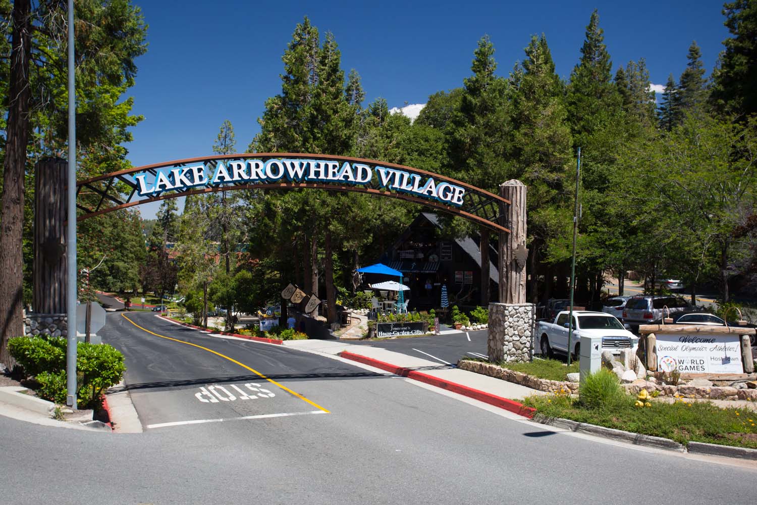 A large wooden archway reading Lake Arrowhead Village stands over a tree-lined road, welcoming visitors to shops and scenic views—perfect for exploring or enjoying Lake Arrowhead, California on a sunny day while participating in the Ho'Omau Altitude Haus multisport training camp.