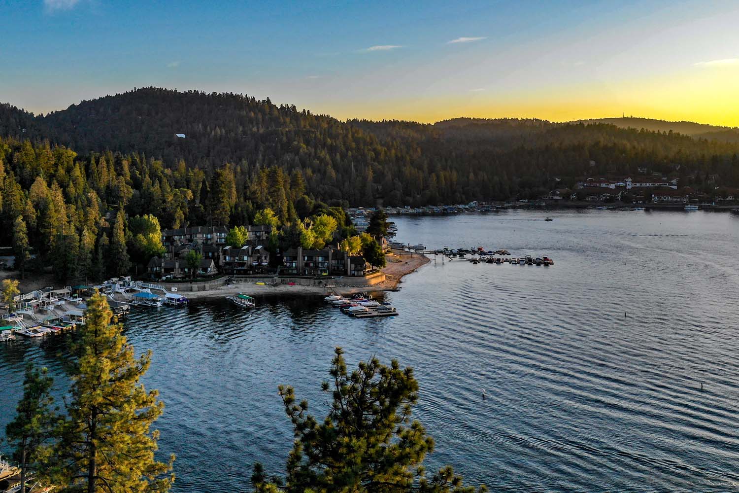 A scenic lake at sunset in Lake Arrowhead, California, surrounded by forested hills. A small marina with boats and a cluster of buildings line the shore, with calm water and golden sunlight. A perfect location for the Ho'Omau Altitude Haus multisport training camp.