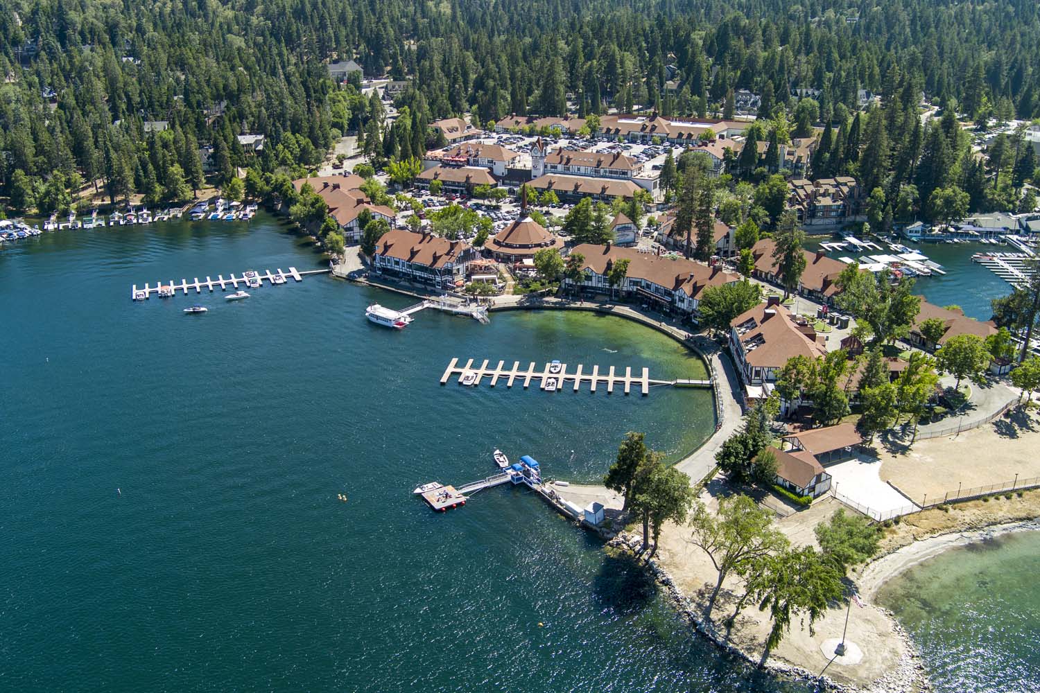 Aerial view of a Lake Arrowhead village with red-roofed buildings surrounded by trees, featuring several boat docks extending into the water and small sandy and grassy areas along the shoreline, an ideal spot for the Ho'Omau Altitude Haus multisport training camp.