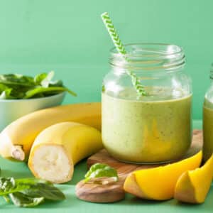 A green smoothie in a glass jar with a striped straw sits on a table beside fresh spinach, banana, and mango slices showcasing a Race-Ready Kitchen mango hulk smoothie recipe.