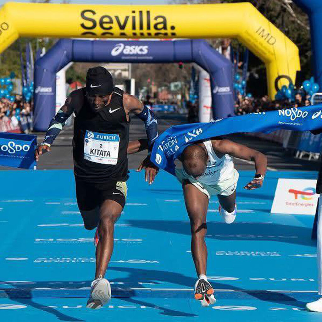 Two male runners cross the finish line at a race in Sevilla—one diving forward to break the tape, the other running upright. The blue finish area is lined with sponsor banners. Featured in Ho'Omau's 'Who Crushed It' weekly global race highlights.