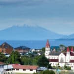 A scenic town with red-roofed buildings and a white church sits by a lake and a snow-capped volcano rising in the background under a partly cloudy sky - setting the stage for Ironman 70.3 Puerto Varas.