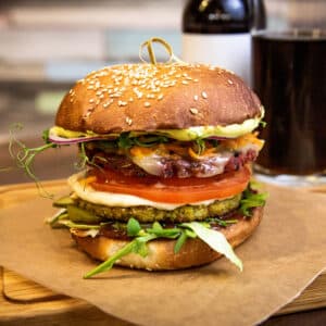 A large, stacked burger with lettuce, tomato, vegan cheese, and a sesame seed bun sits on a wooden board in this Race-Ready Kitchen scene. A glass of dark soda and a bottle are in the background.