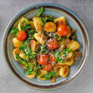 A plate of golden-brown gnocchi with cherry tomatoes, greens, caramelized onions, and grated vegan cheese, viewed from above.