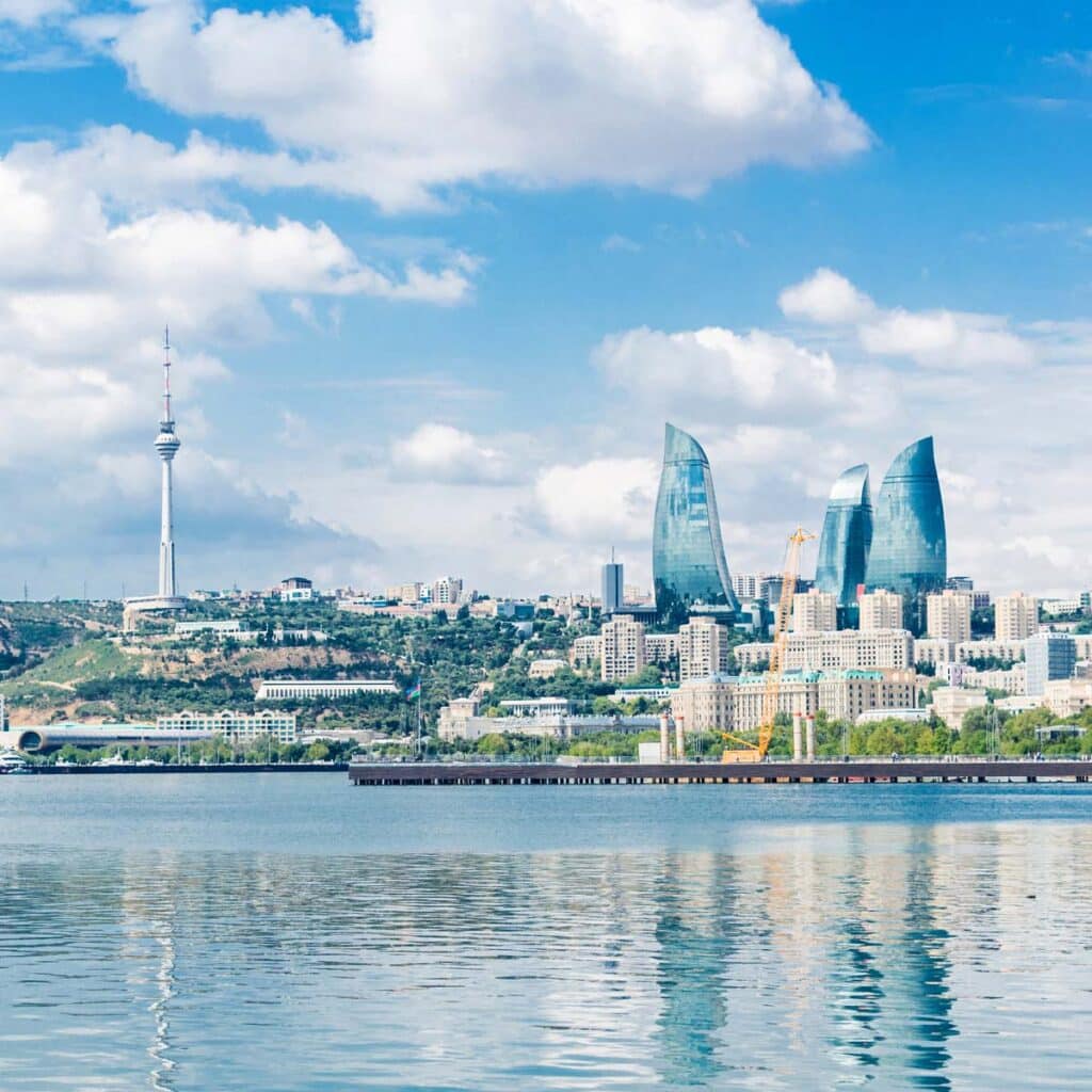 Modern city skyline of Baku, Azerbaijan, featuring the three curved Flame Towers, a tall TV tower, and waterfront buildings reflected on calm water under a partly cloudy sky - setting the stage for Ironman 70.3 Baku.