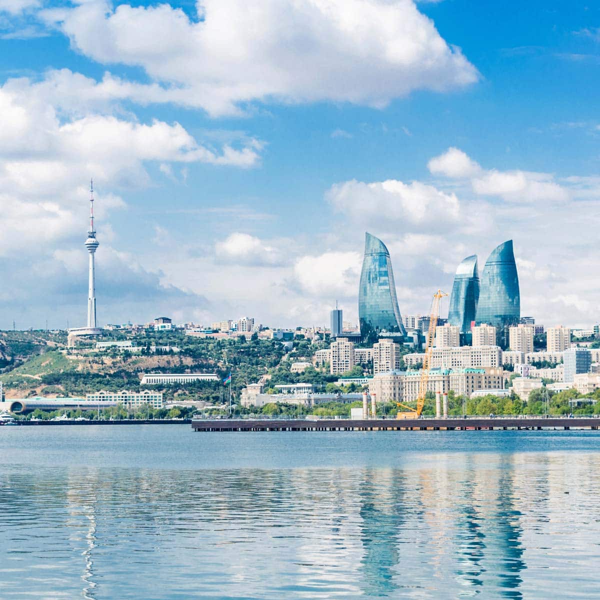 Modern city skyline of Baku, Azerbaijan, featuring the three curved Flame Towers, a tall TV tower, and waterfront buildings reflected on calm water under a partly cloudy sky - setting the stage for Ironman 70.3 Baku.