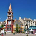 A colorful building with a tall clock tower stands in a lively plaza surrounded by shops, modern architecture, and people under a clear blue sky - setting the stage for Ironman 70.3 Mont Tremblant.