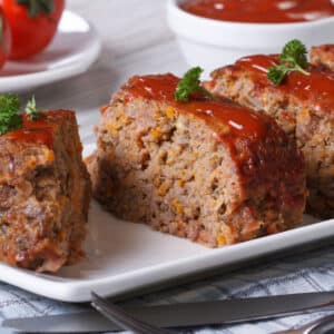Slices of meatloaf topped with a red sauce and garnished with parsley are arranged on a white plate in this Race-Ready Kitchen creation. Fresh tomatoes and a dish of sauce appear in the background.