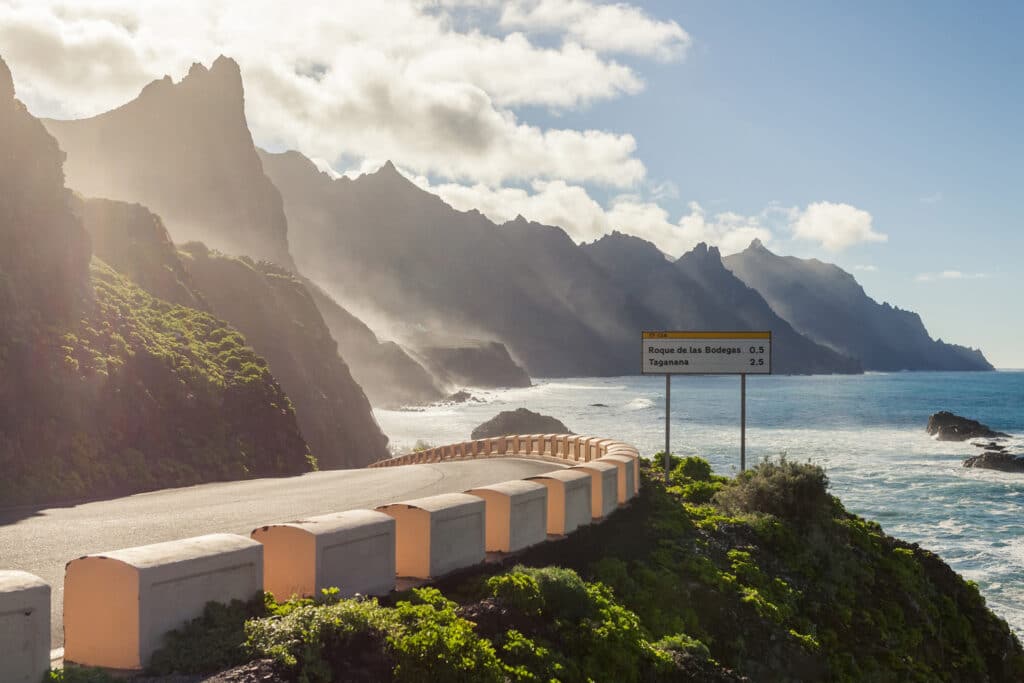 A coastal road curves along dramatic, jagged cliffs with waves crashing below. Sunlight filters through clouds over the mountains as a sign shows distances to nearby locations. A perfect cycling experience for the Ho'Omau Tenerife 'Lava' training camp.