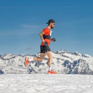 A man wearing a cap, sunglasses, and bright athletic gear runs on snow with snow-capped mountains in the background under a clear blue sky.
