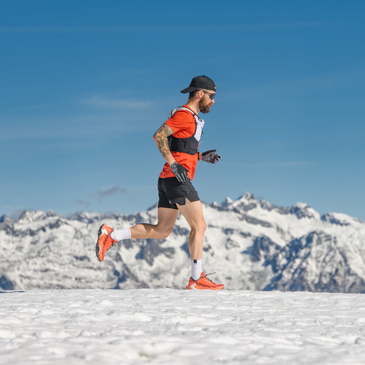 A man wearing a cap, sunglasses, and bright athletic gear runs on snow with snow-capped mountains in the background under a clear blue sky.