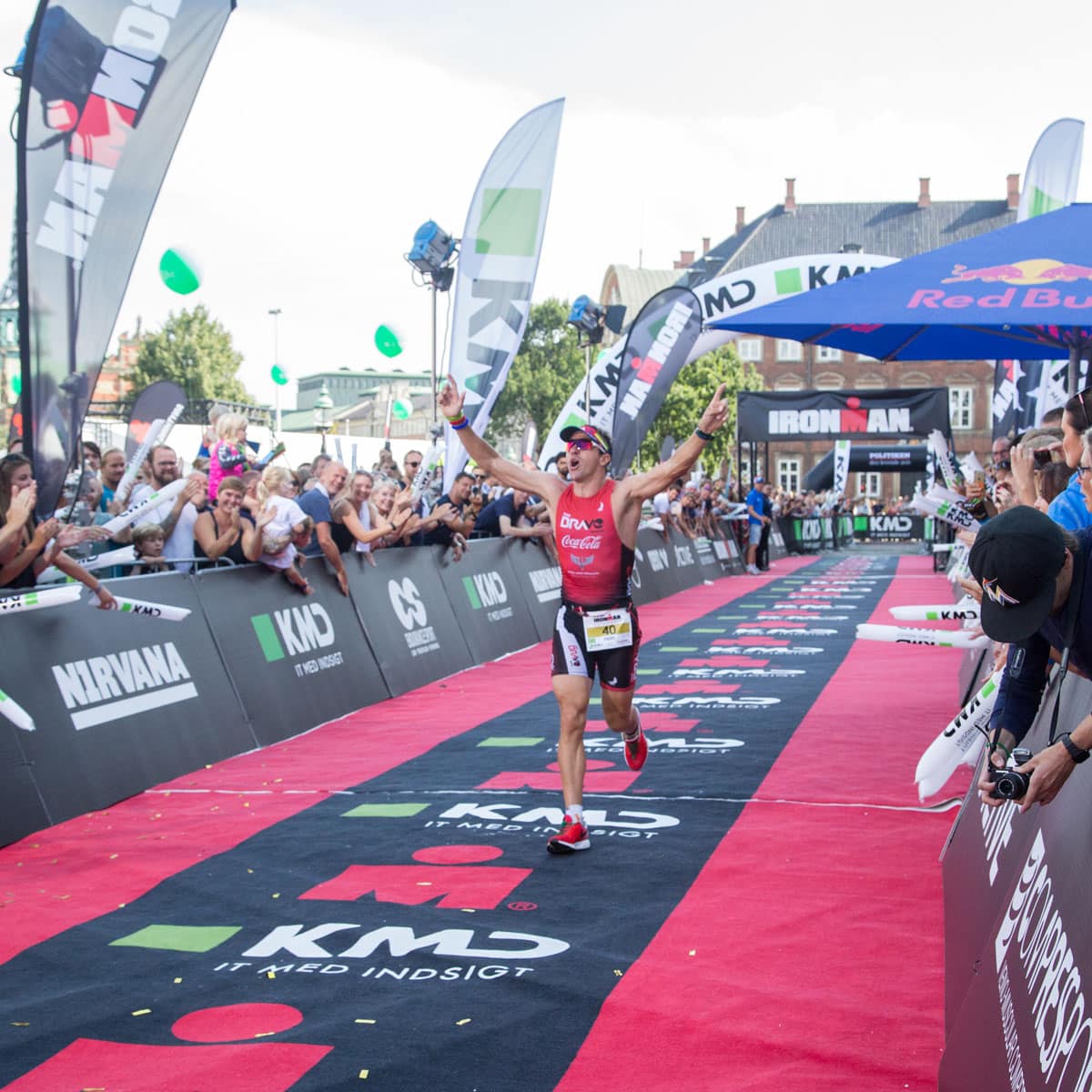 A triathlete wearing a red top and sunglasses crosses the finish line of an Ironman race in under 9 hours, arms raised in victory, as a cheering crowd lines the barriers and banners from sponsors are visible.