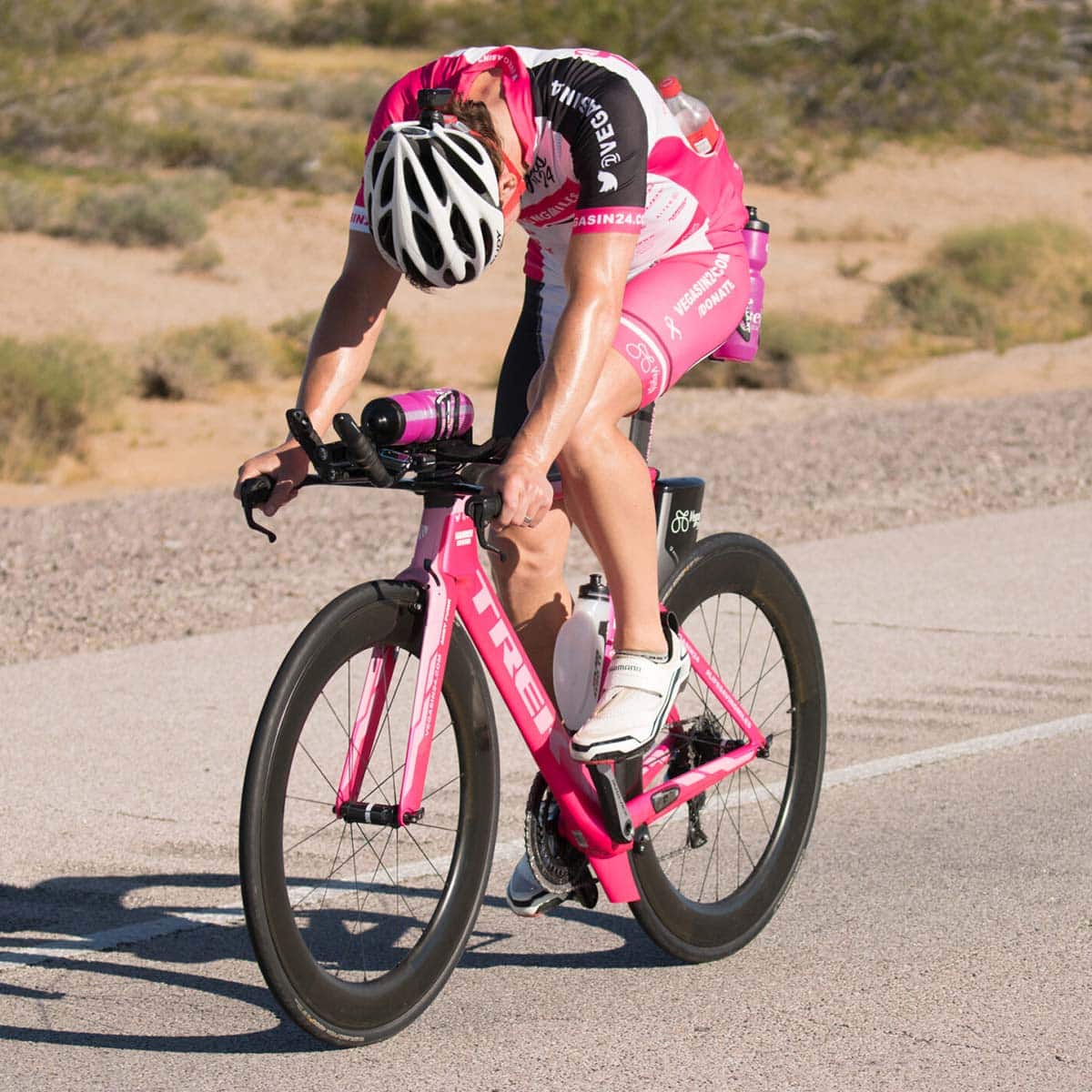 A cyclist in a pink racing outfit and helmet rides a pink bike on a desert road during a Vegas In 24 ultra-cycling challenge, leaning over the handlebars in exhaustion, showcasing the early signs of Vegas Hangover as a result of an extreme long-distance effort.
