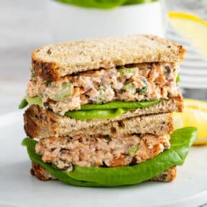 A stack of two plant-based tuna salad sandwiches with lettuce, made with whole grain bread, sits on a white plate next to lemon slices.