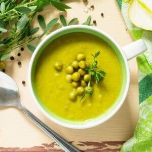 A bowl of green pea soup garnished with peas and herbs sits on a table in a Race-Ready Kitchen setting, surrounded by fresh onion, herbs, a green cloth, and a spoon.