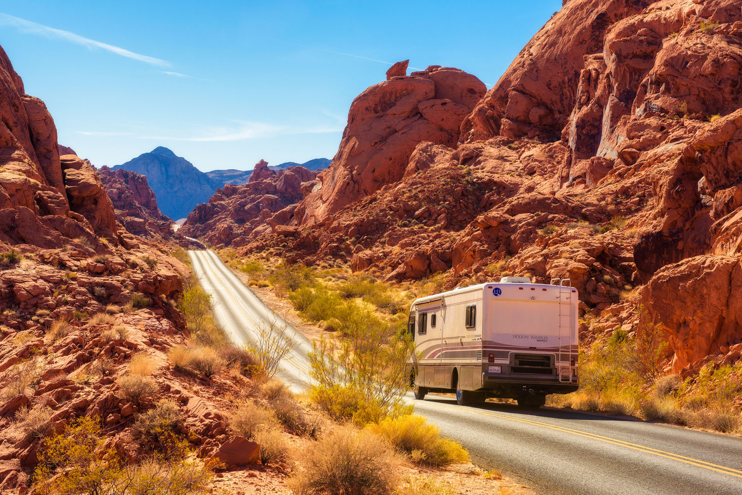 A white RV drives down a winding road through the Valley of Fire, surrounded by red rock formations and sparse desert vegetation under a bright blue sky.