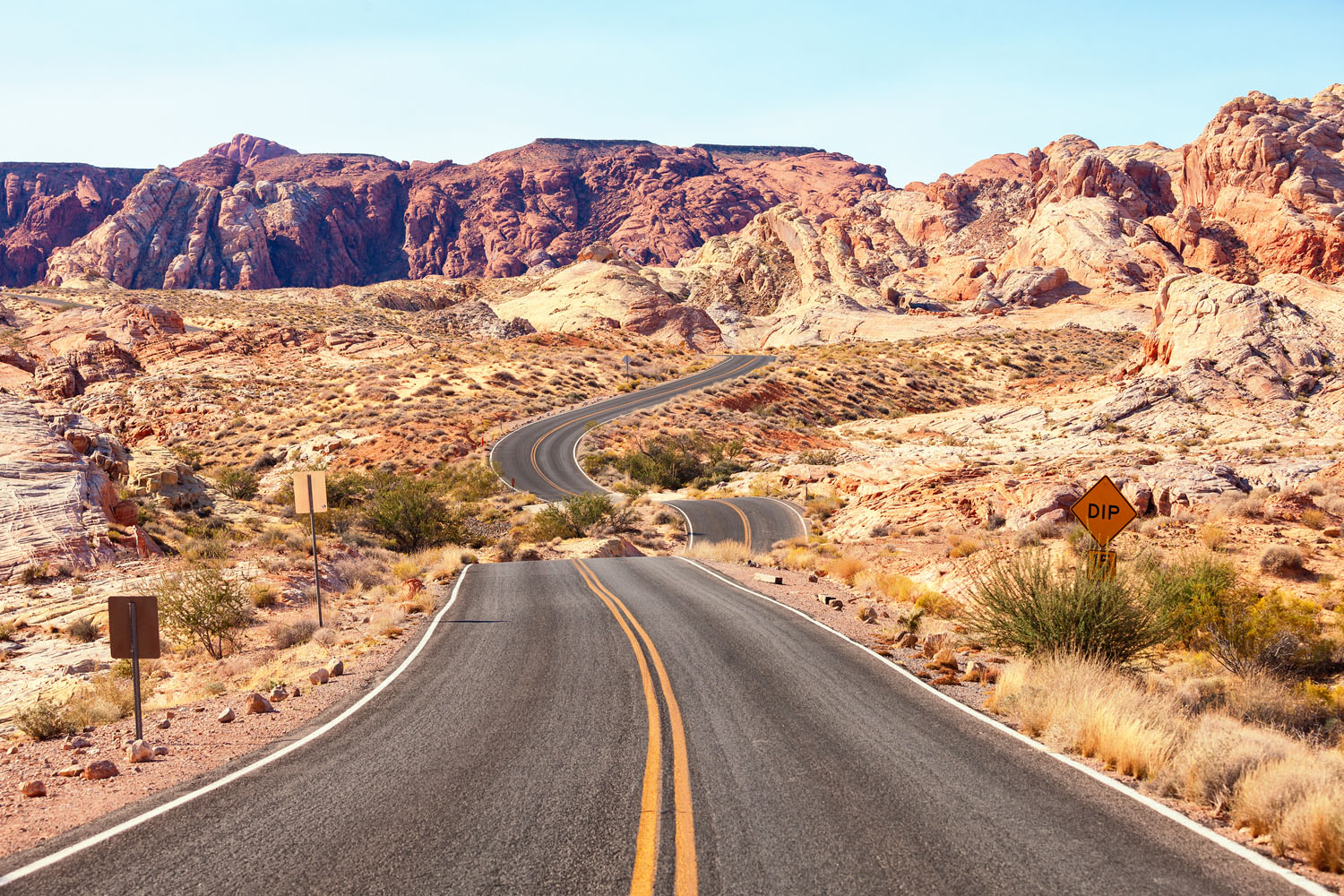 A winding road stretches through the dry, rocky Valley of Fire desert landscape with red and tan mountains in the background under a clear blue sky; a yellow Dip road sign is visible on the right.