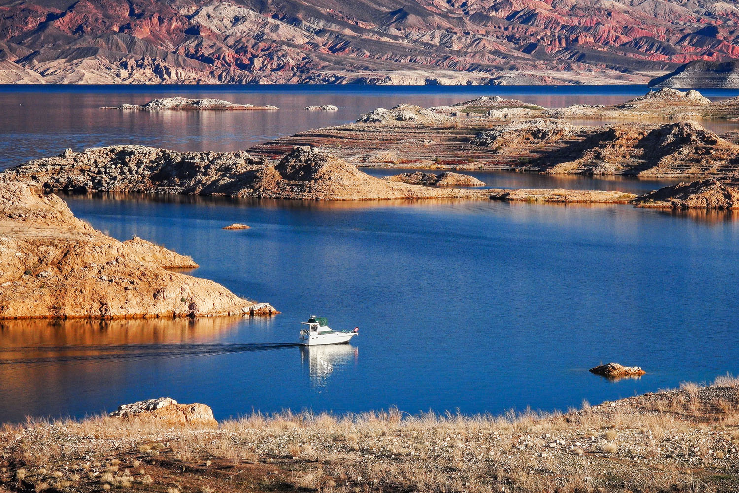 A small white boat sails on Lake Mead near the Valley of Fire RV Training Camp, surrounded by rocky, arid islands and rugged, colorful hills in the background under clear skies.
