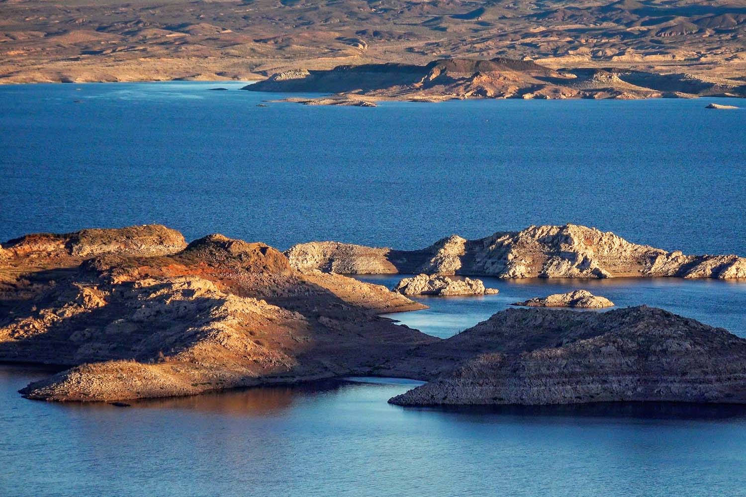 A wide expanse of deep blue lake water surrounds several rocky, sunlit islands with rugged terrain, near the Valley of Fire RV Training Camp, set against a dry, brown landscape in the background under a clear sky.