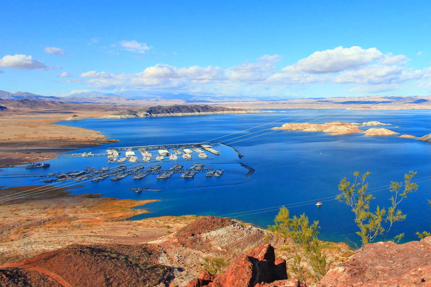 Lake Mead with several boat docks is surrounded by an arid, rocky landscape near the Valley of Fire RV Training Camp, under a bright sky with scattered clouds. Sparse vegetation and distant mountains complete the scenic backdrop.