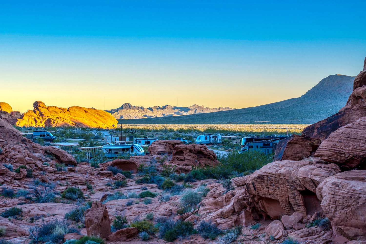 A scenic view of the Valley of Fire RV Training Camp nestled among red rock formations in a desert landscape, with mountains in the distance under a clear blue sky at sunrise or sunset.