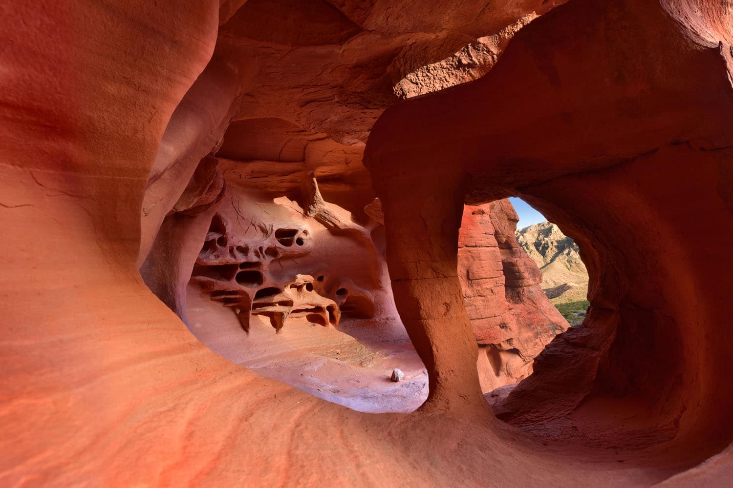 View from inside a red sandstone cave in Valley of Fire, with smooth, curving walls and honeycomb-like formations, looking out through a large round opening onto sunlit desert hills and blue sky.