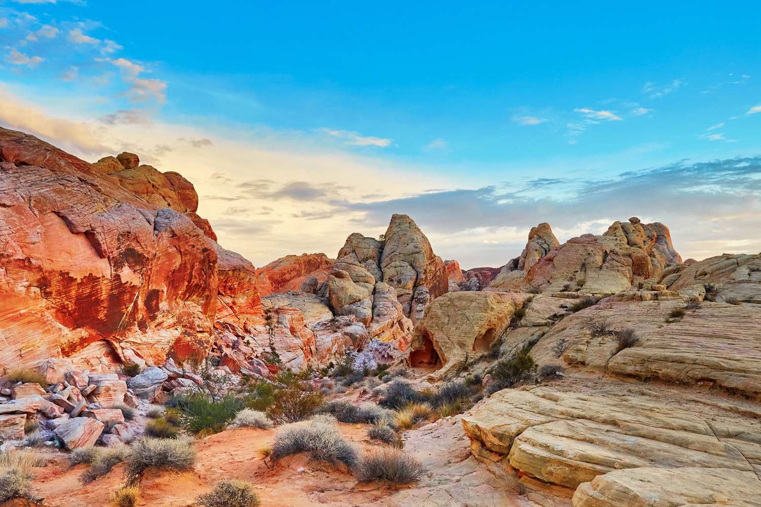 Colorful desert landscape at Valley of Fire RV Training Camp, featuring orange, red, and tan rock formations, scattered shrubs, and a bright blue sky with some clouds overhead.