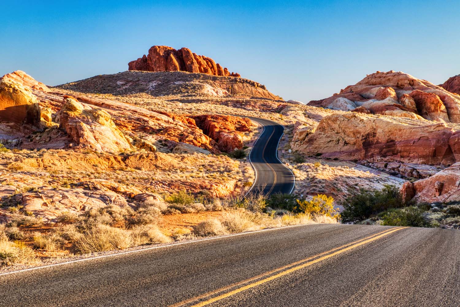 A winding road curves through colorful desert rock formations at the Valley of Fire RV Training Camp, with sunlight casting warm tones on the rugged landscape beneath a clear blue sky.