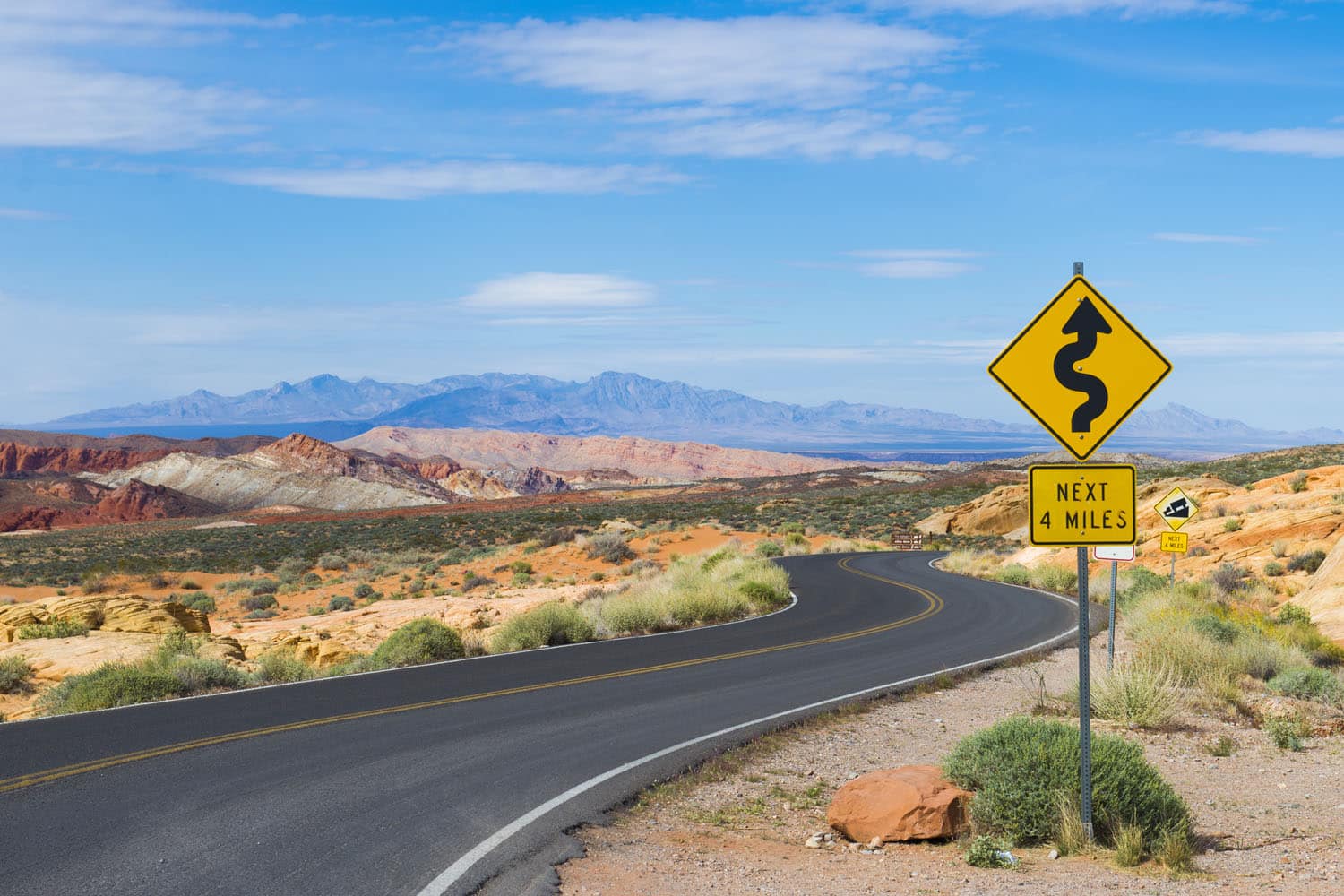 A winding road curves through the Valley of Fire’s desert landscape, flanked by colorful rocks and distant mountains under a blue sky. A yellow road sign warns of curves for the next 4 miles amid sparse roadside vegetation.