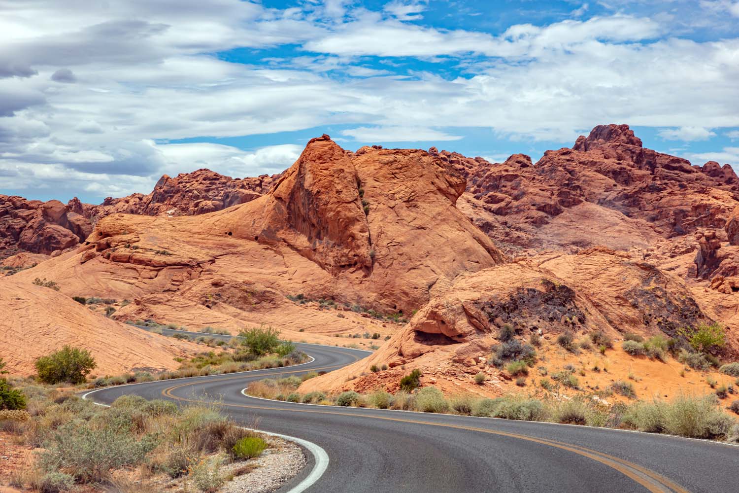 A winding road curves through the Valley of Fire, showcasing orange, rocky formations and sparse vegetation under a partly cloudy blue sky.