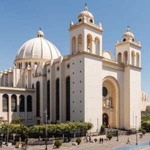 A large white cathedral in San Salvador with a dome and bell towers stands against a clear blue sky. Green trees and people are visible in the foreground.