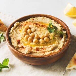 A bowl of hummus topped with chickpeas, olive oil, parsley, and paprika sits on a table with pita bread, lemon wedges, and fresh parsley.