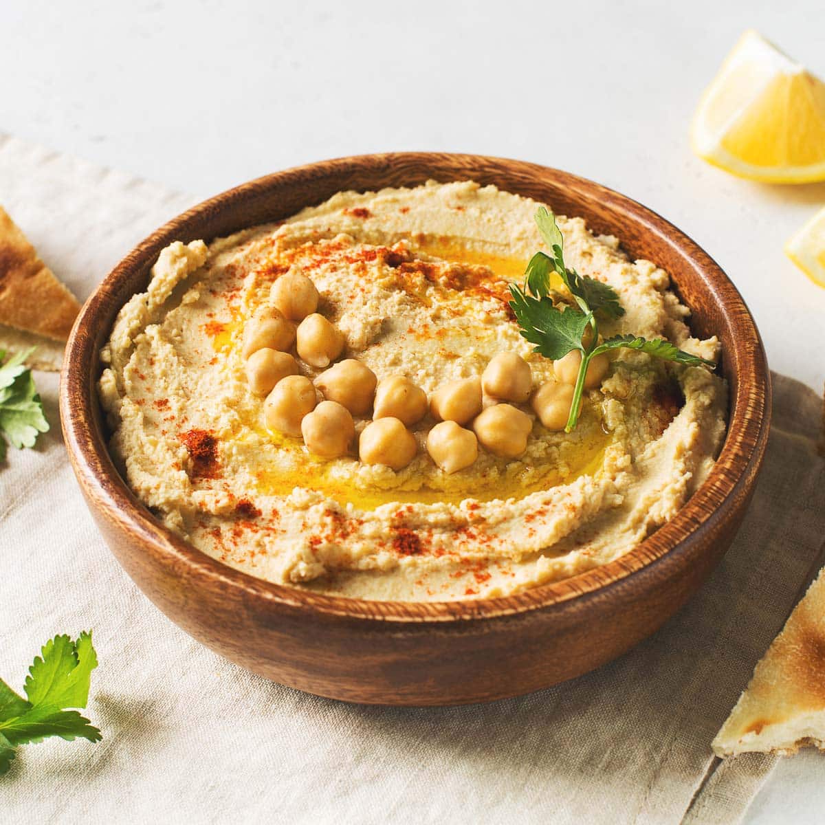 A bowl of hummus topped with chickpeas, olive oil, parsley, and paprika sits on a table with pita bread, lemon wedges, and fresh parsley.