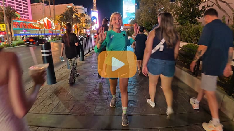 Jacqueline Busse in a green shirt smiles while walking on the Las Vegas strip at night, discussing Ho'Omau's endurance sports crossword challenge, surrounded by other people and bright neon lights.