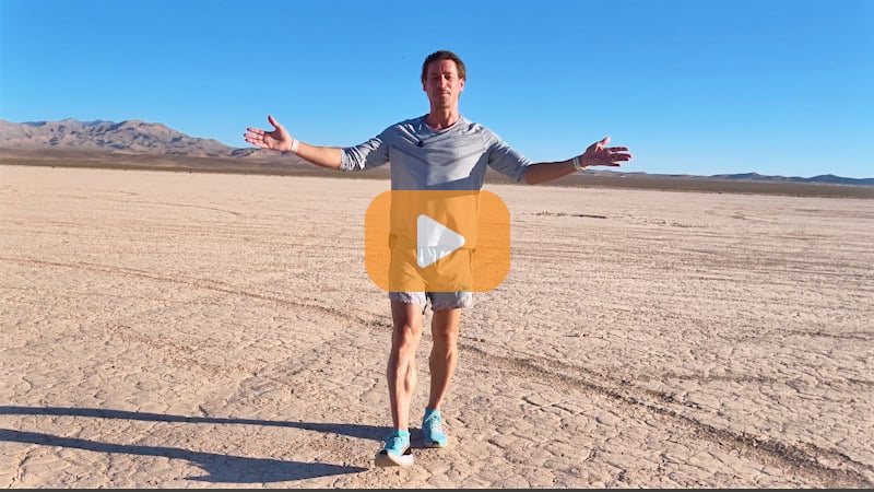 Andy Funk in athletic clothing with arms outstretched stands on a dry, cracked desert landscape under a clear blue sky, evoking the spirit of adventure found along long distance cycling routes, with distant mountains in the background.