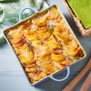A square blue baking dish filled with golden-brown vegan scalloped potatoes garnished with rosemary sits on a wooden table beside a green napkin and a basket with green fabric—perfect for any Race-Ready Kitchen.