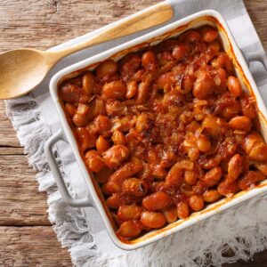 A white baking dish filled with baked beans in sauce sits on a wooden table with a wooden spoon and a gray cloth, ready to fuel your Race-Ready Kitchen.