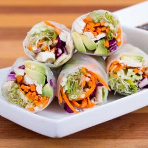 Fresh spring rolls filled with tofu, avocado, shredded carrots, cabbage, and sprouts are stacked on a white plate beside a bowl of dipping sauce. Race-Ready Kitchen recipes ensure you enjoy vibrant flavors with every bite.